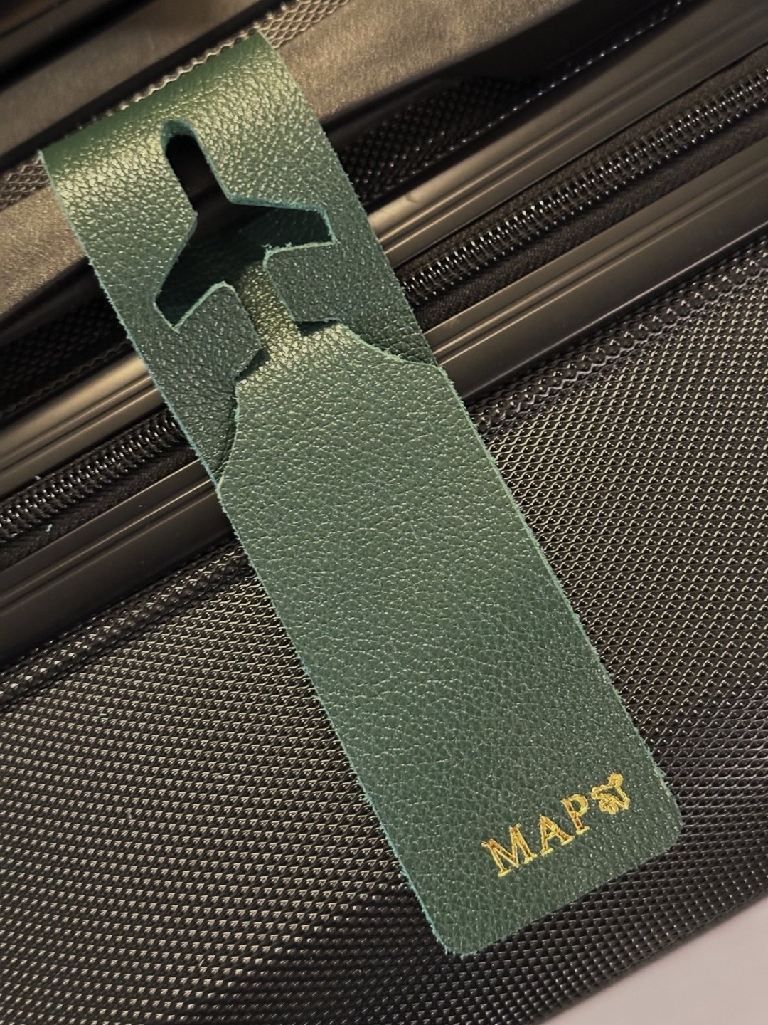 Tag de mala em couro verde com recorte de avião e monograma dourado, sobre mala preta